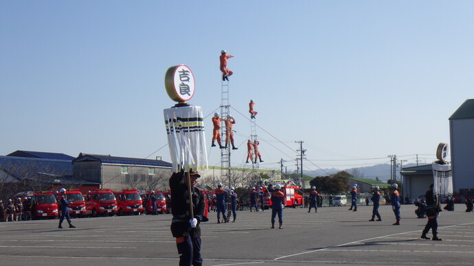 三連はしごの上で演技をする消防職員と纏を振る消防団員