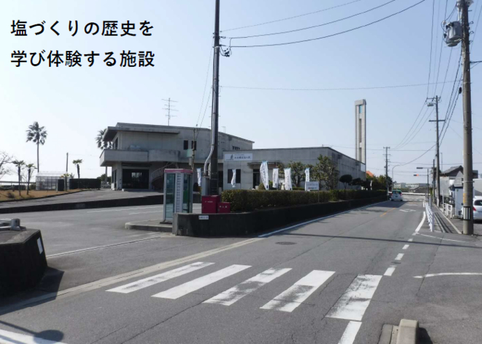 塩田体験館 駐車場の入り口写真（入り口から続くアスファルト駐車場の奥に建物が見える）